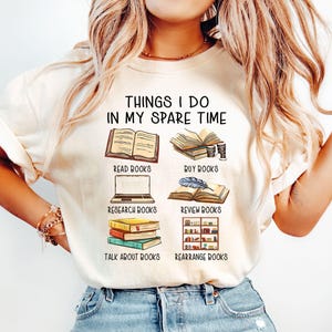 Può includere: T-shirt color crema con la scritta "THINGS I DO IN MY SPARE TIME" e illustrazioni di libri. La maglietta presenta immagini che rappresentano la lettura, l'acquisto, la ricerca, la revisione, la conversazione e la riorganizzazione di libri.