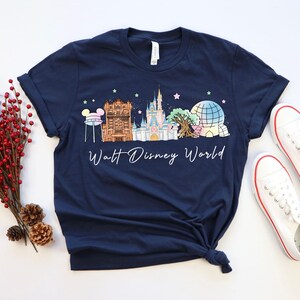 May include: Navy blue t-shirt featuring a colorful illustration of Walt Disney World landmarks. The design includes a castle, Spaceship Earth, and other iconic structures. The words "Walt Disney World" are written in a cursive font. White sneakers with red trim are next to the shirt.