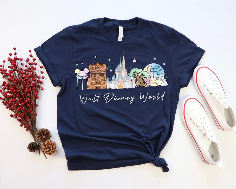 May include: Navy blue t-shirt featuring a colorful illustration of Walt Disney World landmarks. The design includes a castle, Spaceship Earth, and other iconic structures. The words "Walt Disney World" are written in a cursive font. White sneakers with red trim are next to the shirt.
