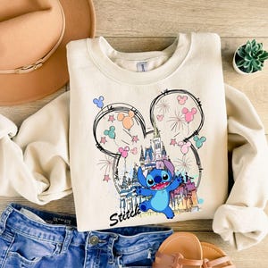 May include: Cream-colored sweatshirt featuring a colorful graphic of Stitch from Lilo & Stitch, with a castle, fireworks, and Mickey Mouse ears. The word "Stitch" is written in a script font. The sweatshirt is paired with blue jeans and sandals.
