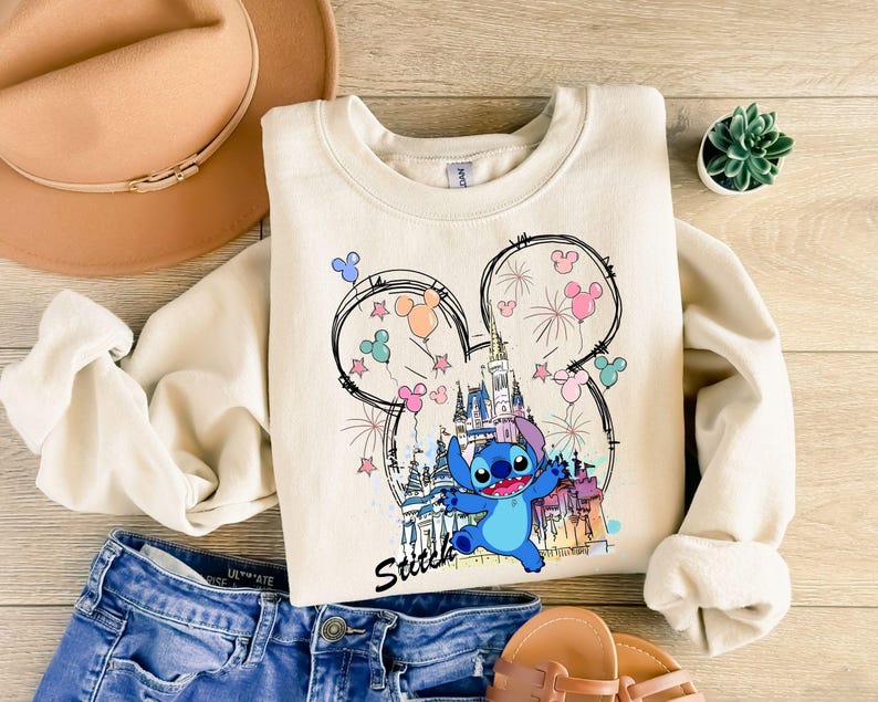 May include: Cream-colored sweatshirt featuring a colorful graphic of Stitch from Lilo & Stitch, with a castle, fireworks, and Mickey Mouse ears. The word "Stitch" is written in a script font. The sweatshirt is paired with blue jeans and sandals.