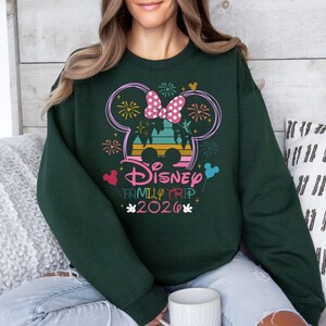 May include: Dark green sweatshirt featuring a colorful Disney Family Trip 2020 design. The design includes a Mickey Mouse silhouette with fireworks, a bow, and the text "Disney Family Trip 2020" in a playful font. The sweatshirt is a comfortable, casual style.