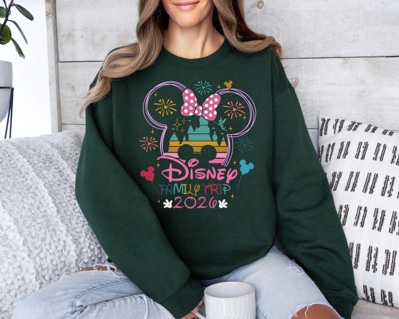May include: Dark green sweatshirt featuring a colorful Disney Family Trip 2020 design. The design includes a Mickey Mouse silhouette with fireworks, a bow, and the text "Disney Family Trip 2020" in a playful font. The sweatshirt is a comfortable, casual style.
