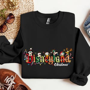 May include: Black crewneck sweatshirt with the words "Disneyland Christmas" in colorful lettering. The design includes cartoon characters and festive holiday decorations. The sweatshirt is made of a soft material.