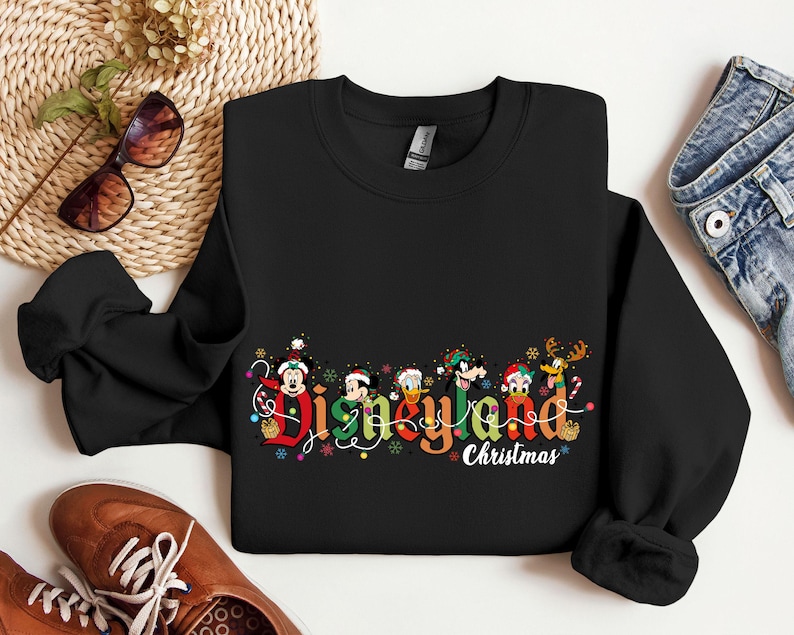 May include: Black crewneck sweatshirt with the words "Disneyland Christmas" in colorful lettering. The design includes cartoon characters and festive holiday decorations. The sweatshirt is made of a soft material.
