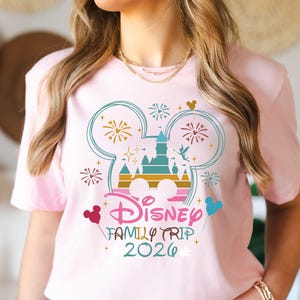 May include: A light pink t-shirt featuring a colorful Disney design. The design includes a castle, fireworks, and the text "Disney Family Trip 2026" in a playful font. Mickey Mouse ears and balloons are also included in the design.