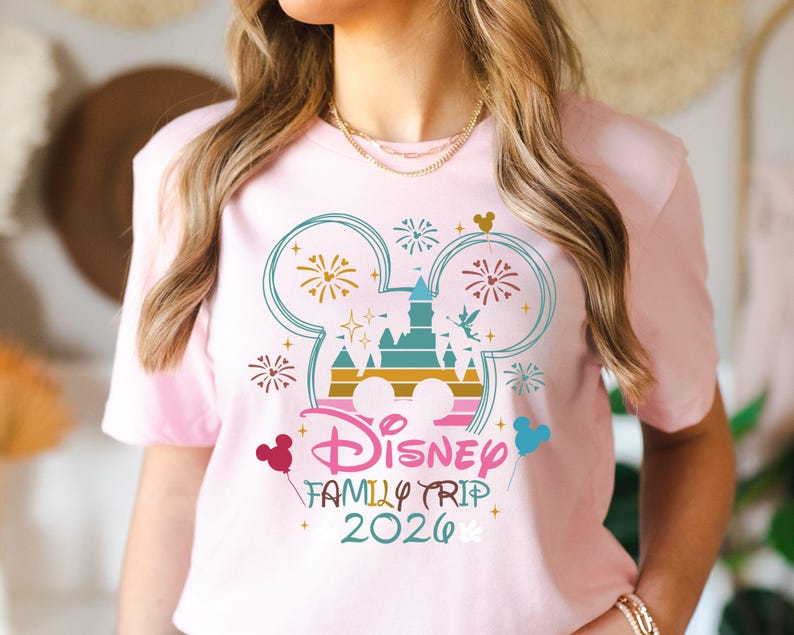 May include: A light pink t-shirt featuring a colorful Disney design. The design includes a castle, fireworks, and the text "Disney Family Trip 2026" in a playful font. Mickey Mouse ears and balloons are also included in the design.