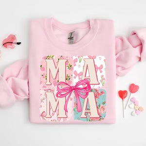 May include: A light pink sweatshirt with the word "MAMA" in a floral and bow design. The letters are in a light beige color, with a large pink bow in the center. The sweatshirt is surrounded by heart-shaped sunglasses and candy.