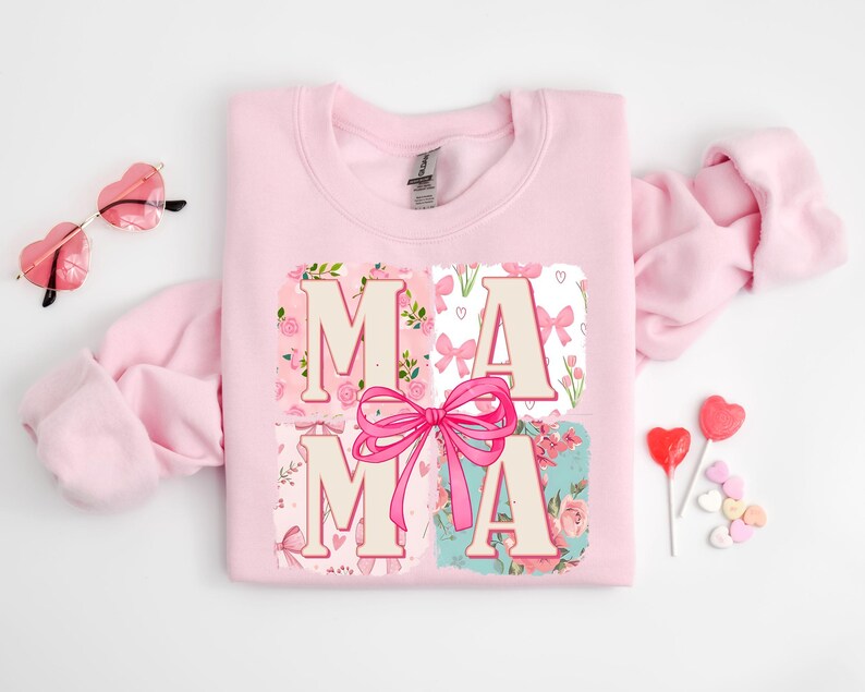May include: A light pink sweatshirt with the word "MAMA" in a floral and bow design. The letters are in a light beige color, with a large pink bow in the center. The sweatshirt is surrounded by heart-shaped sunglasses and candy.
