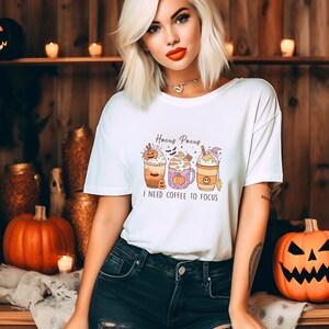May include: White t-shirt with a Halloween-themed design featuring three coffee cups with pumpkin and witch hat decorations. The text "Hocus Pocus I need coffee to focus" is printed on the shirt.