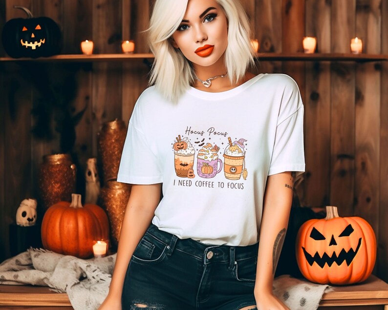 May include: White t-shirt with a Halloween-themed design featuring three coffee cups with pumpkin and witch hat decorations. The text "Hocus Pocus I need coffee to focus" is printed on the shirt.