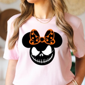 Puede incluir: Camiseta rosa claro con un dise&ntilde;o gr&aacute;fico negro de una cara sonriente y un lazo naranja con lunares. La cara del personaje tiene ojos blancos y una boca cosida. La camiseta es de cuello redondo.