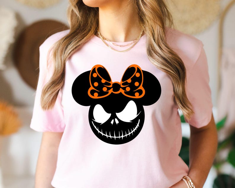 Puede incluir: Camiseta rosa claro con un dise&ntilde;o gr&aacute;fico negro de una cara sonriente y un lazo naranja con lunares. La cara del personaje tiene ojos blancos y una boca cosida. La camiseta es de cuello redondo.