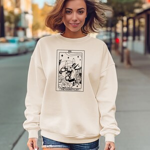 Pode incluir: Uma camisa de moletom cor creme com um design de carta de tarô em preto e branco que apresenta um par de tesouras e o texto "The Hairstylist".