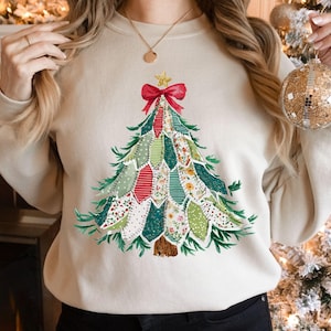 以下が含まれることがあります： クリーム色のスウェットシャツ。クリスマスツリーのデザイン。ツリーは緑、赤、花柄の布パッチで構成され、赤いリボンと金の星が飾られています。お祝いのホリデーアパレルアイテムです。
