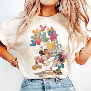 Könnte beinhalten: Cremefarbenes T-Shirt mit einem Vintage-Grafik von Mickey und Minnie Mouse. Mickey hält bunte Luftballons, und Minnie hält einen Sonnenschirm. Ein Schloss ist im Hintergrund. Das Design hat eine Retro-Cartoon-Ästhetik.