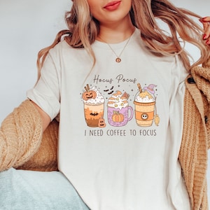 May include: A white t-shirt with a graphic of three pumpkin spice coffee drinks. The text "Hocus Pocus I need coffee to focus" is printed on the shirt.