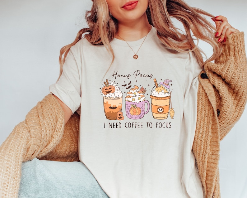 May include: A white t-shirt with a graphic of three pumpkin spice coffee drinks. The text "Hocus Pocus I need coffee to focus" is printed on the shirt.