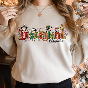 May include: Beige sweatshirt with the words "Disneyland Christmas" in colorful lettering. The word "Disneyland" is decorated with Mickey Mouse, Minnie Mouse, Donald Duck, Goofy, and other Disney characters. The sweatshirt is a festive holiday item.