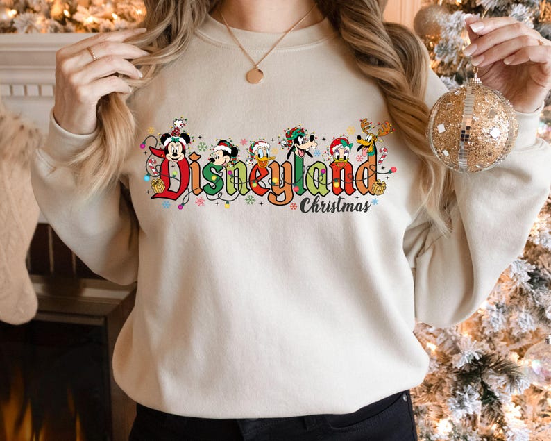 May include: Beige sweatshirt with the words "Disneyland Christmas" in colorful lettering. The word "Disneyland" is decorated with Mickey Mouse, Minnie Mouse, Donald Duck, Goofy, and other Disney characters. The sweatshirt is a festive holiday item.