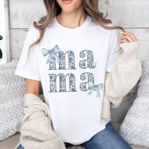 K&ouml;nnte beinhalten: Wei&szlig;es T-Shirt mit dem Wort "mama" in blau-wei&szlig;em Blumenmuster bedruckt. Die Buchstaben sind mit blauen Schleifen verziert.