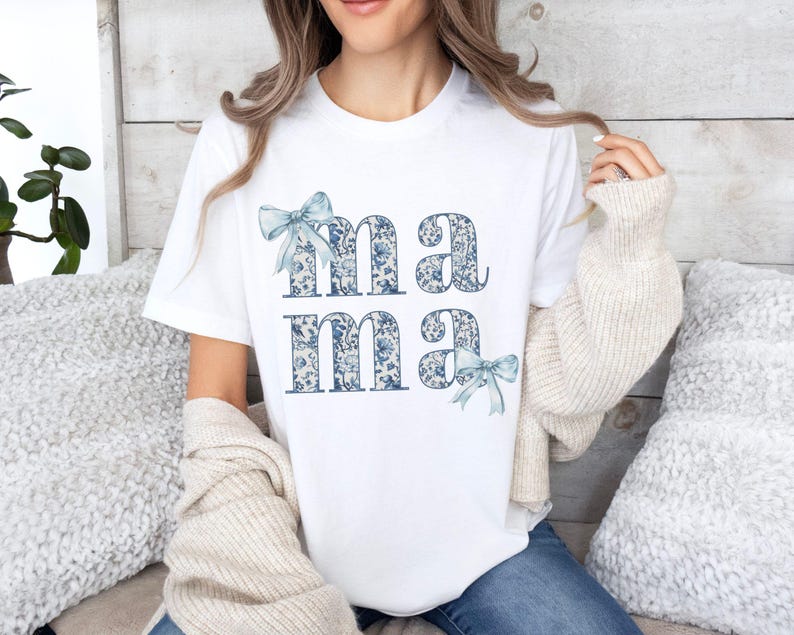 K&ouml;nnte beinhalten: Wei&szlig;es T-Shirt mit dem Wort "mama" in blau-wei&szlig;em Blumenmuster bedruckt. Die Buchstaben sind mit blauen Schleifen verziert.