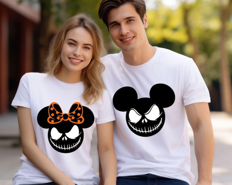 Puede incluir: Dos camisetas blancas con un dise&ntilde;o gr&aacute;fico negro y naranja. Una camiseta presenta un dise&ntilde;o inspirado en Mickey Mouse con un lazo, y la otra tiene un dise&ntilde;o similar sin lazo. El gr&aacute;fico tiene un tema espeluznante de Halloween.