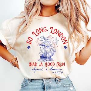 May include: Cream-colored t-shirt with a nautical design. The graphic features a blue and red sailing ship, fireworks, and text that reads "So Long, London, Had a Good Run, Signed, America, 1776."