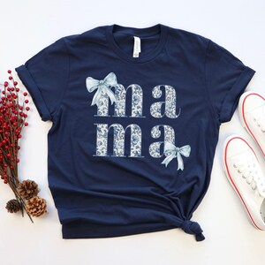 K&ouml;nnte beinhalten: Ein marineblaues T-Shirt mit dem Wort "mama" in wei&szlig; mit einem blauen Blumenmuster bedruckt. Die Buchstaben sind mit blauen Schleifen verziert.