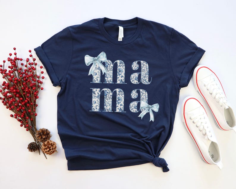 K&ouml;nnte beinhalten: Ein marineblaues T-Shirt mit dem Wort "mama" in wei&szlig; mit einem blauen Blumenmuster bedruckt. Die Buchstaben sind mit blauen Schleifen verziert.