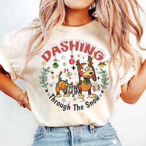 May include: A white t-shirt with a Christmas-themed design featuring a cartoon dog wearing a Santa hat and surrounded by lights, candy canes, and snowflakes. The text "Dashing Through The Snow" is printed below the dog.