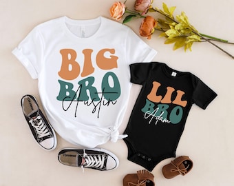 Puede incluir: Dos camisetas con el texto "Big Bro Austin" y "Lil Bro Adam" en una fuente retro con colores naranja, verde y blanco. Las camisetas están sobre una superficie marrón claro.