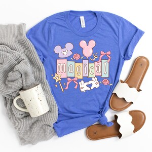 May include: A blue t-shirt with the word "Magical" in pastel colors, featuring cartoon Mickey Mouse ears, bows, and a magic wand. A gray sweater, a speckled mug, and white sandals with brown soles are also in the image.