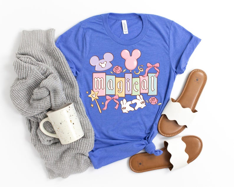 May include: A blue t-shirt with the word "Magical" in pastel colors, featuring cartoon Mickey Mouse ears, bows, and a magic wand. A gray sweater, a speckled mug, and white sandals with brown soles are also in the image.