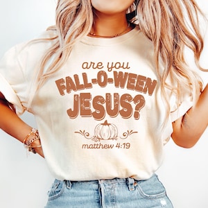 Puede incluir: Camiseta color crema con el texto "are you FALL-O-WEEN JESUS?" en letras marrones. Un gráfico de calabaza está centrado debajo, con el versículo bíblico Mateo 4:19. La camiseta es un diseño casual con temática de otoño.