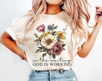 May include: Cream-colored t-shirt featuring a floral bouquet in shades of pink, yellow, blue, and white. The design includes green leaves and the text "in the waiting GOD IS WORKING" in a script font. The shirt has rolled-up sleeves.