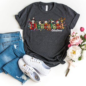 May include: Dark gray t-shirt with the words "Disneyland Christmas" in a colorful design featuring Mickey Mouse, Donald Duck, and other Disney characters. The shirt is paired with blue jeans and white sneakers.