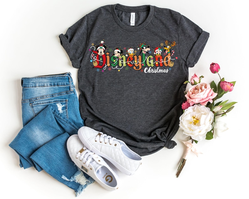 May include: Dark gray t-shirt with the words "Disneyland Christmas" in a colorful design featuring Mickey Mouse, Donald Duck, and other Disney characters. The shirt is paired with blue jeans and white sneakers.