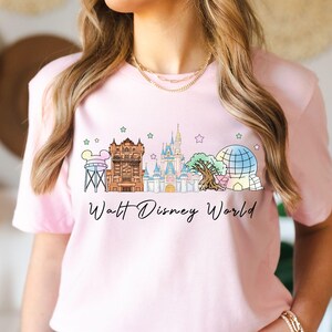 May include: A light pink t-shirt featuring a colorful graphic of Walt Disney World landmarks, including Cinderella Castle and Spaceship Earth. The words "Walt Disney World" are written in a script font below the graphic.