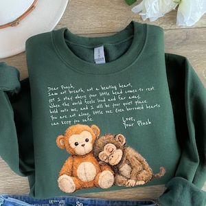 May include: Dark green sweatshirt featuring a graphic of two plush monkeys and handwritten text. The monkeys are brown and tan, with one resting its head on the other. The text reads "Dear Punch, I am not breath...Love, Your Plush."