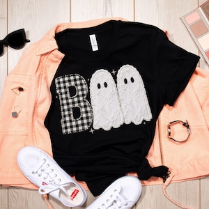 May include: A black t-shirt featuring the word BOO in a graphic design. The letter B is a black and white buffalo check pattern, while the two Os are white lace ghosts. The shirt is layered under a peach-coloured jacket with white trainers, sunglasses, a makeup palette, and jewellery.