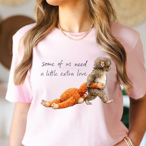 Peut inclure: T-shirt rose clair avec l'inscription "some of us need a little extra love". L'illustration repr&eacute;sente un singe portant un jouet en peluche orange. Le singe est gris et marron. Le t-shirt est &agrave; manches courtes.