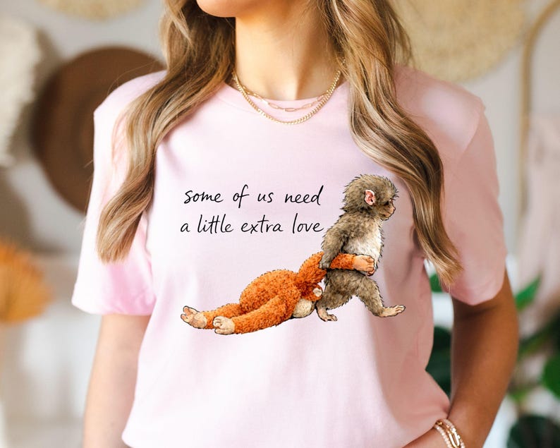 Peut inclure: T-shirt rose clair avec l'inscription "some of us need a little extra love". L'illustration repr&eacute;sente un singe portant un jouet en peluche orange. Le singe est gris et marron. Le t-shirt est &agrave; manches courtes.