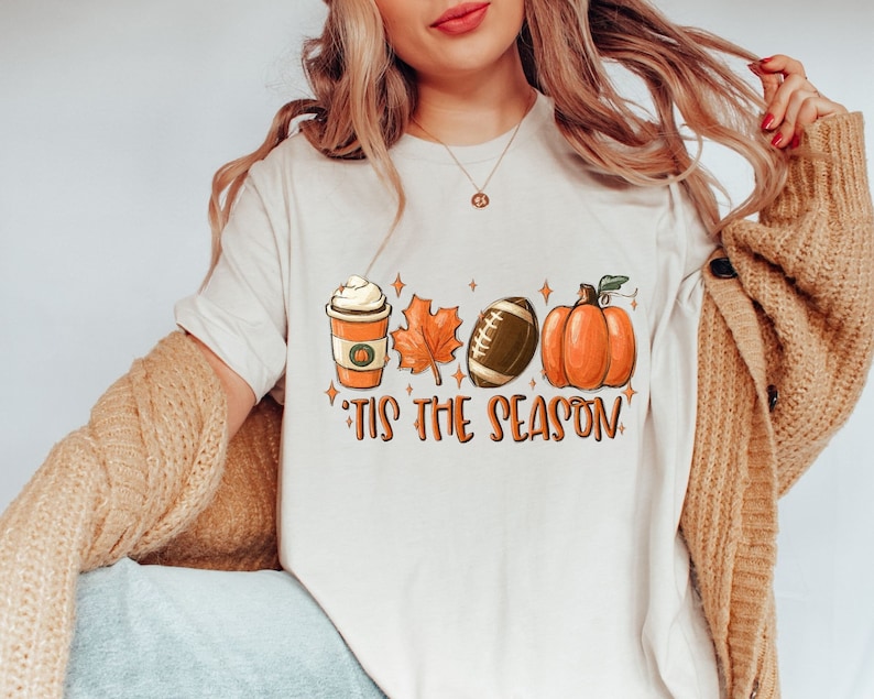 K&ouml;nnte beinhalten: Wei&szlig;es T-Shirt mit einem Aufdruck einer K&uuml;rbis, einem Football, einem Ahornblatt und einer Tasse Kaffee mit Schlagsahne. Der Text "'Tis the season" befindet sich unter dem Aufdruck.