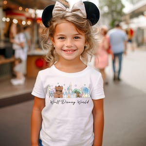 May include: A young girl wearing a white t-shirt with a colorful graphic of Walt Disney World landmarks. The shirt features a castle, a globe, and other iconic structures. She is also wearing a black and gold Minnie Mouse ear headband.