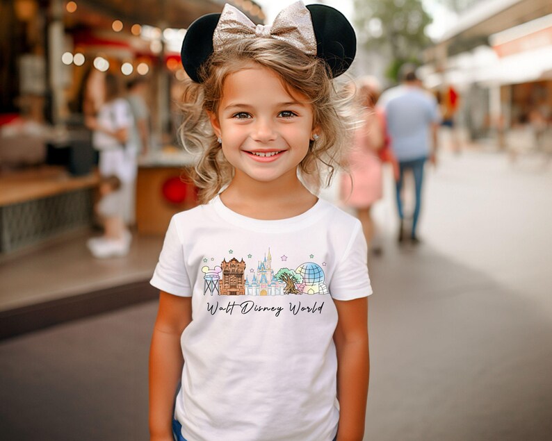 May include: A young girl wearing a white t-shirt with a colorful graphic of Walt Disney World landmarks. The shirt features a castle, a globe, and other iconic structures. She is also wearing a black and gold Minnie Mouse ear headband.