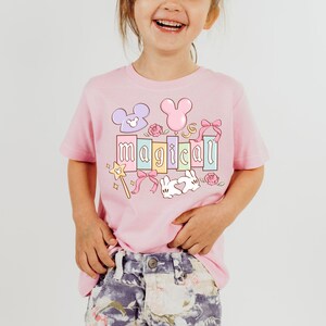 May include: A light pink t-shirt with the word "magical" in pastel colors. The design includes a Mickey Mouse head, a bunny, a wand, and other decorative elements. The shirt is worn by a young child.