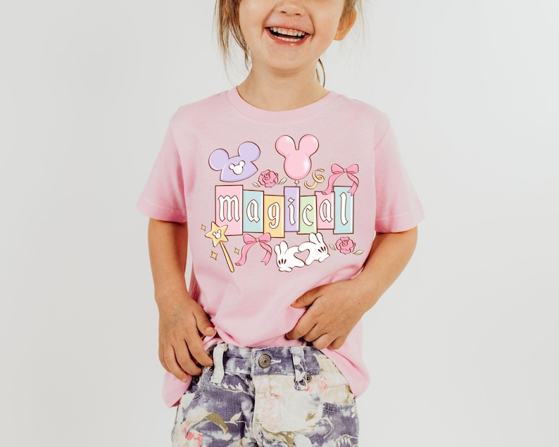 May include: A light pink t-shirt with the word "magical" in pastel colors. The design includes a Mickey Mouse head, a bunny, a wand, and other decorative elements. The shirt is worn by a young child.