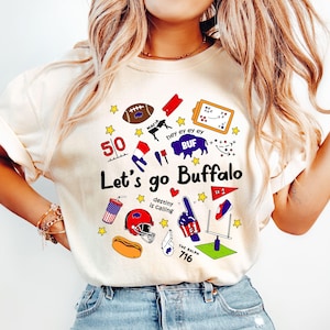 May include: Cream-colored t-shirt with a graphic design featuring football-themed illustrations and the text "Let's go Buffalo." The design includes a football, helmet, and the number 716.