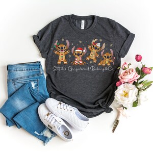 Puede incluir: Camiseta gris oscuro con cuatro personajes de dibujos animados de pan de jengibre, adornados con accesorios festivos. El texto "Stitch's Gingerbread Baking & Co" est&aacute; impreso debajo. La camiseta se combina con vaqueros azules y zapatillas blancas.